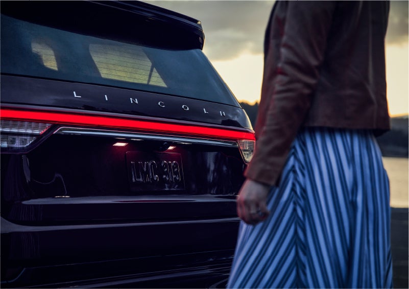 A person is shown near the rear of a 2023 Lincoln Aviator® SUV as the Lincoln Embrace illuminates the rear lights | Lidtke Lincoln in Beaver Dam WI