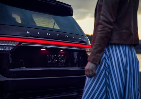 A person is shown near the rear of a 2024 Lincoln Aviator® SUV as the Lincoln Embrace illuminates the rear lights | Lidtke Lincoln in Beaver Dam WI